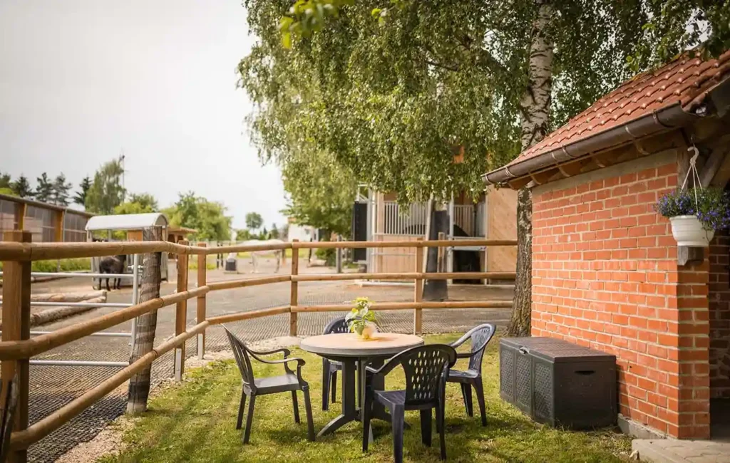 Ein kleiner runder Gartentisch mit vier Stühlen steht auf einer grünen Rasenfläche. Rechts daneben ist ein Teil eines Gebäudes zu sehen, umgeben von einem Zaun. Im Hintergrund befinden sich Stallbereiche mit Pferdeboxen und Pferden, die dort fressen. Die Szene wirkt ruhig und einladend, wie ein gemütlicher Platz zum Verweilen in der Nähe des Pferdestalls.