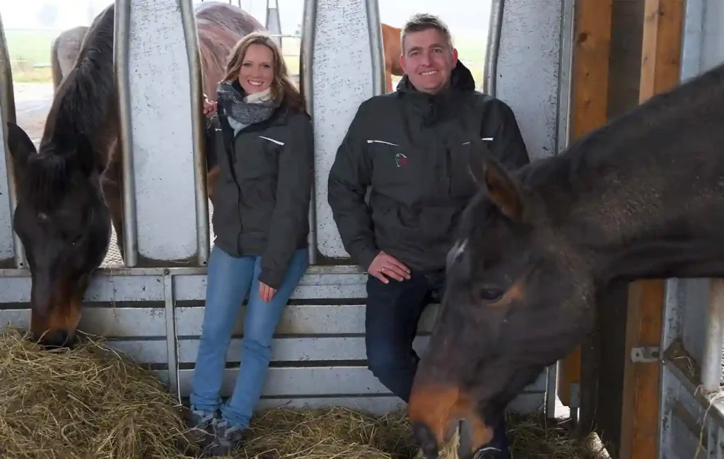 Das Bild zeigt das Innere einer Pferdebox. Durch zwei getrennte Öffnungen stecken draußen stehende Pferde ihre Köpfe hinein, um Heu zu fressen. In der Box sitzt eine Frau auf dem Rand der linken Öffnung und ein Mann auf dem Rand der rechten Öffnung. Beide lächeln in die Kamera, während die Pferde aus den neben ihnen befindlichen Öffnungen ruhig fressen.