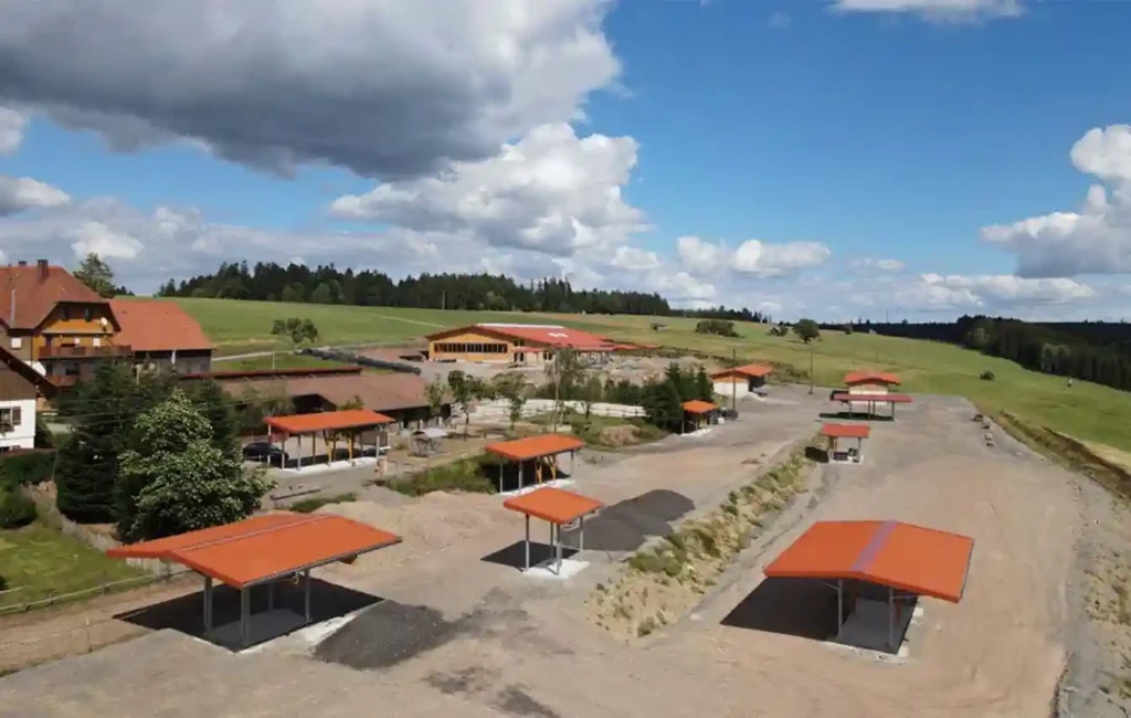 Das Bild zeigt ein Hofgelände aus der Vogelperspektive. Mehrere kleine Überdachungen oder offene Unterstände stehen verteilt auf dem Gelände. Links am Rand befindet sich ein größeres Haus. Das gesamte Areal ist von grüner Wiese und Bäumen umgeben und liegt an einem leichten Abhang. Die Aufnahme vermittelt einen umfassenden Überblick über den Hof und seine Außenbereiche.