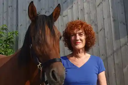 Eine Frau schaut lächelnd in die Kamera. Links neben ihr steht ein braunes Pferd. Von beiden sind nur Kopf und Oberkörper sichtbar. Im Hintergrund befindet sich ein graues Gebäude.