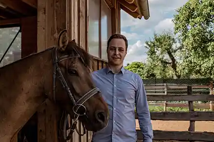 Ein Mann steht neben einem braunen Pferd und schaut in die Kamera. Das Pferd steht ruhig an seiner Seite. Im Hintergrund befinden sich von links aus gesehen ein braunes Gebäude, ein Zaun und mehrere Bäume.