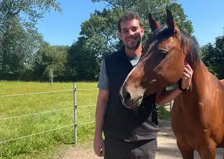 In der Bildmitte steht ein lächelnder Mann, der direkt in die Kamera schaut und seinen Arm unterhalb des Kopfes eines braunen Pferdes hält, das rechts neben ihm steht. Hinter ihnen liegt eine grüne Wiese, die durch einen Zaun vom sandigen Untergrund getrennt ist, auf dem der Mann steht.