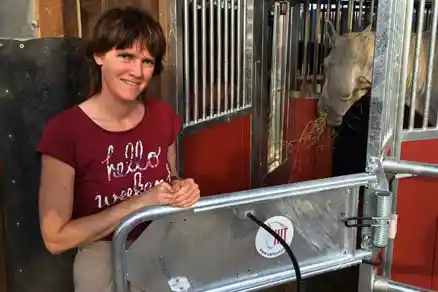 Das Bild zeigt eine Frau, die in der Öffnung einer Pferdebox steht, lächelt und in die Kamera schaut. Rechts neben ihr, innerhalb der Box, steht ein Pferd, das Heu frisst.