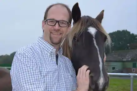 Ein Mann hält den Kopf eines schwarzen Pferdes fest, welches rechts von ihm positioniert ist und lächelt dabei. Von beiden ist jeweils nur der Kopf zu sehen. Im Hintergrund sind eine Scheune und eine Wiese leicht zu erkennen, dominierend ist jedoch der helle Himmel.