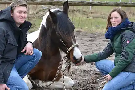 Das Bild bildet ab, wie ein Mann und eine Frau auf einem sandigen, erdigen Untergrund hocken und lächelnd in die Kamera schauen. Zwischen ihnen kniet ein Pferd bzw. legt sich entspannt auf dem Boden ab. Die Szene wirkt warm, freundlich und vertraut.