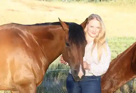 Das Bild zeigt eine Frau, die auf einer Wiese steht und in die Kamera schaut. Sie hält den Kopf eines braunen Pferdes, das links neben ihr steht. Die Szene wirkt ruhig und natürlich.