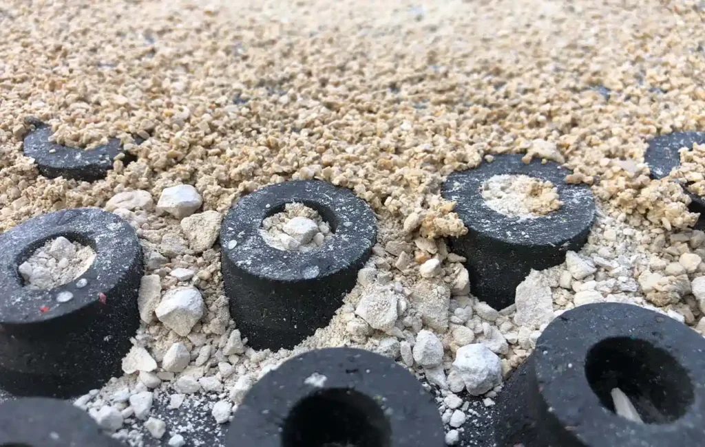 Zu sehen ist eine Nahaufnahme eines Bodenaufbaus. Im Vordergrund sieht man mehrere runde, schwarze Gummielemente mit löchriger Mitte. Darüber liegt eine Schicht aus hell-beigem körnigem Material, das wie Sand-Kies-Gemisch oder Reitplatzsand mit Zuschlagstoffen aussieht. Das Material wirkt locker und trocken.