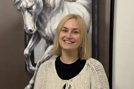 Eine Frau lächelt und schaut direkt in die Kamera. Hinter ihr hängt ein Bild, das ein Pferd zeigt.