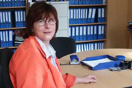 Eine Frau sitzt links im Bild an ihrem Arbeitsplatz und schaut in die Kamera. Hinter ihr stehen mehrere Regale, die dicht mit Ordnern gefüllt sind.