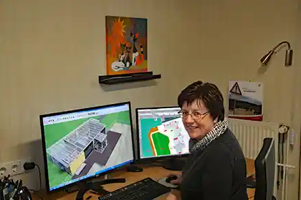 Das Bild zeigt eine Frau, die von hinten zu sehen ist und sich über die Schulter zur Kamera umdreht. Sie sitzt an einem Computerarbeitsplatz.